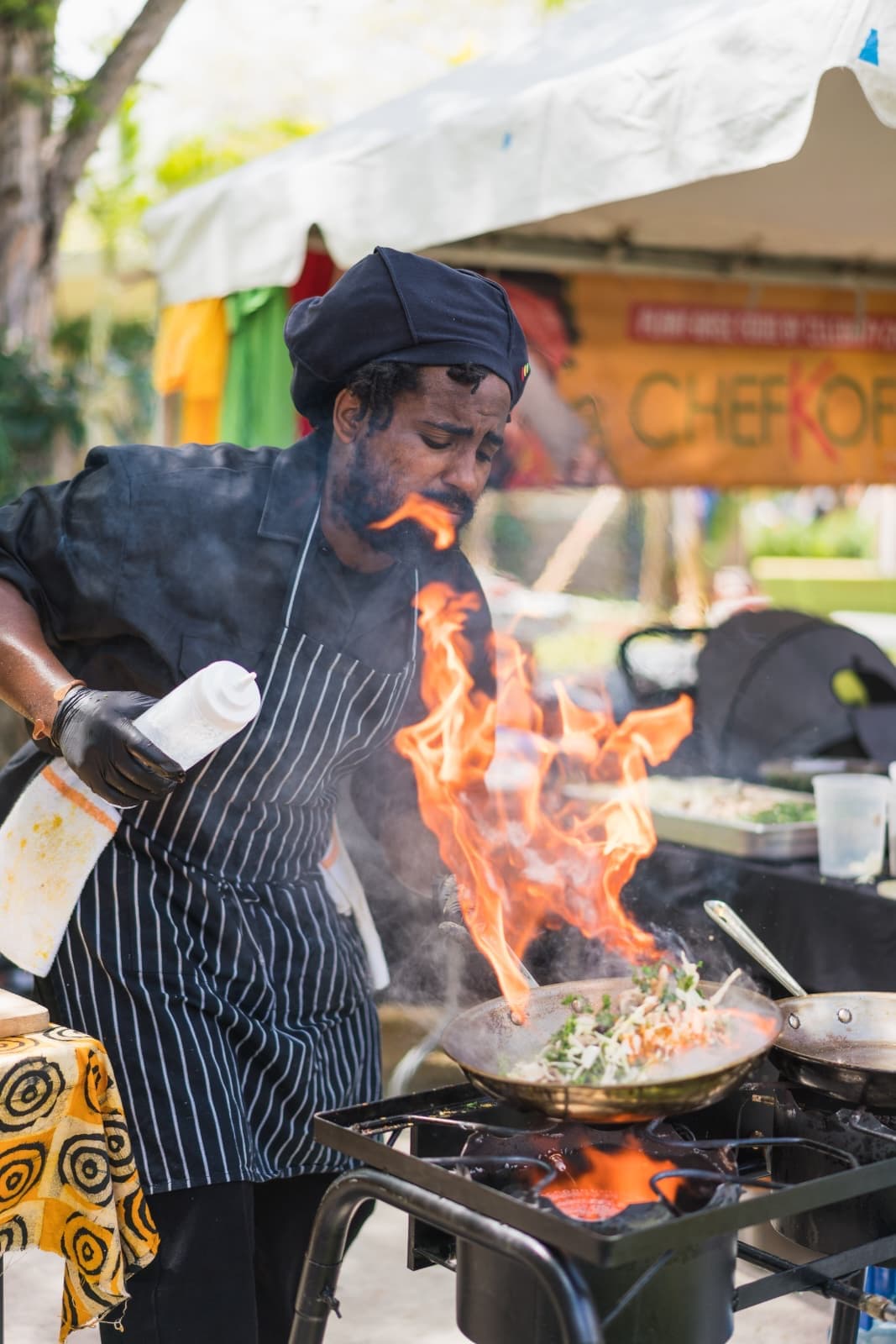Chef Kriss Kofi flambéing a wok over open flame at a live event